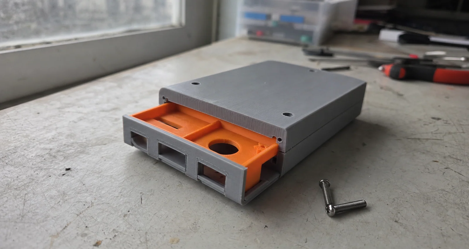 Gray PLA electronics enclosure prototype with an orange fit-check insert on a used workshop bench